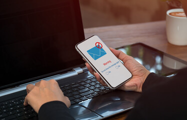 A businesswoman works on a laptop computer on a table, while a warning window appears on the screen of her cell phone. Spam e-mail blocking, phishing email warning pop-up, network security concept