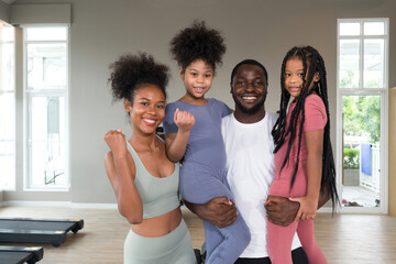 Happy family enjoy holiday together in fitness center. Young father hold both of his daughter in arms while the mother raise her fist up.