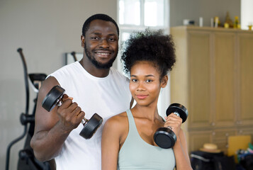 Young curly black hair couple enjoy holiday together in fitness center.  Both lifting barbell with one hand.
