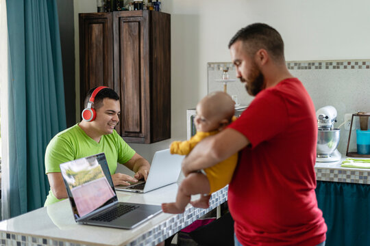 Image of a family of two gay men with a foster baby using technology in their home, baby being held by a bearded man looking at a computer and her husband sitting using his laptop with headphones.
