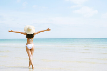 backside young woman skin tan in sunhat and bikini standing with her arms raised to her head enjoying on the beach vacation travel. and enjoy life at sea looking view of beach ocean on hot summer day.