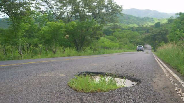 LOW ANGLE, CLOSE UP, DOF: Pick-up truck drives along a crumbling country road in lush green central Panama. Old pickup truck drives past a large pothole filled with murky water. Decaying gravel road.