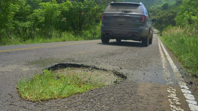 SLOW MOTION, LOW ANGLE, CLOSE UP, DOF: Modern SUV rental car drives into a massive water filled pothole in the middle of an asphalt road crossing the vibrant exotic countryside in central Panama.