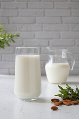 A glass of almond milk next to almonds and sprigs of greenery, in the background a decanter of milk.