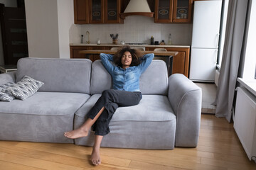 Carefree sleepy Black teen girl relaxing on soft couch, stretching body, taking deep breath of...