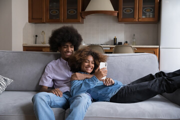 Cheerful dating teen Black couple using online app on smartphone at home. Happy young African...