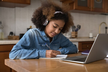 Serious busy high school student teenage girl in wireless headphones studying at home, writing notes, sitting at kitchen table, using laptop, watching video class, learning webinar