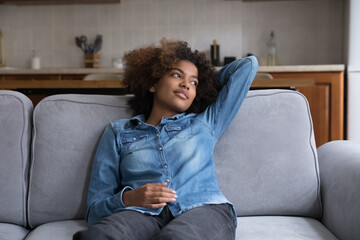 Thoughtful positive Black teen girl relaxing on comfortable sofa in living room, taking deep breath of fresh air, enjoying break, leisure time at home, looking away, thinking, dreaming