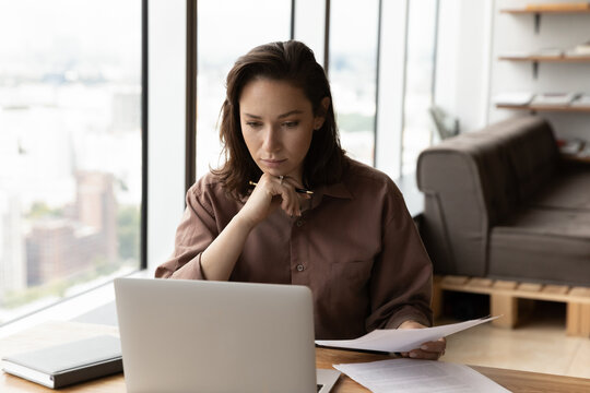 Pensive Business Professional Checking Documents At Workplace, Using Laptop, Consulting Online Legal Service On Internet, Holding Papers, Reading Text On Screen. Businesswoman Doing Paperwork