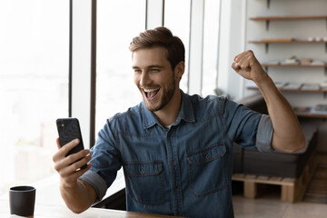 Happy millennial man in casual excited with good news, using smartphone, looking at screen, making...