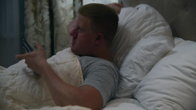 Caucasian man in grey t-shirt lies in bed with his smart phone at night. Man lies under blanket on white pillow. Real time video. Social media addiction theme.