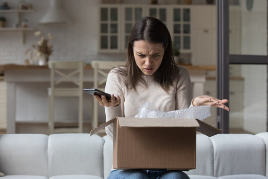 Puzzled dissatisfied customer getting damaged good from Internet store, opening wrong parcel, finding mistake. Confused worried young woman holding smartphone, checking carton box