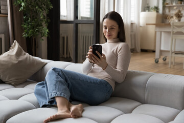 Obraz premium Positive focused young cellphone user woman shopping on Internet on mobile phone at home, chatting online, using virtual app on smartphone, resting, sitting on couch, enjoying leisure