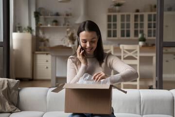 Happy satisfied Internet shop customer girl unwrapping parcel at home, making telephone call to...