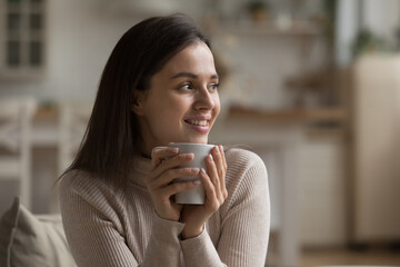 Cheerful dreamy girl holding mug of hot beverage, looking away in deep good thoughts, smiling,...