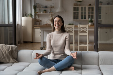 Happy cheerful young yogi woman practicing meditation on couch, sitting in lotus pose, keeping zen...