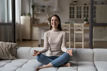 Fotobehang Happy cheerful young yogi woman practicing meditation on couch, sitting in lotus pose, keeping zen hands, fingers, laughing, smiling, looking at camera. Yoga, mindfulness concept. Indoor portrait © fizkes