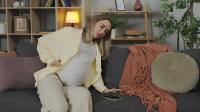 Exhausted expectant mother in last month of pregnancy suffering from backache at home. Pregnant woman with severe pain massaging her lumbar muscles while sitting on sofa.