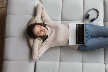 Carefree peaceful pretty 20s girl relaxing on soft cozy couch near big wireless headphones,...