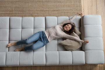 Happy peaceful young woman relaxing on comfortable couch at home with closed eyes, stretching hands...