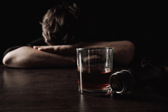 Man with glass and bottle of whiskey lying or sleeping on table at night. Alcohol addiction concept