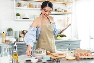 young Asian housewife in kitchen using tablet for searching recipes online cooking the bakery dough homemade.
