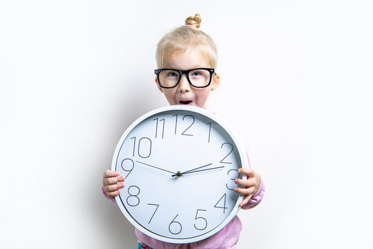 Surprised child girl in glasses holding a large wall clock on a light background.