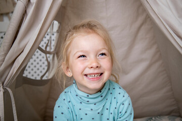Smiling blonde child girl in play tent or wigwam at home