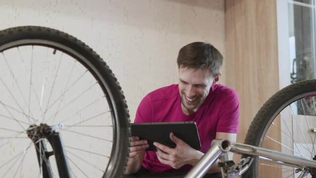 Man with tablet sits near inverted bicycle with pedals removed. Guy taps screen and joyful, happy with smile looks at bike in room. Cyclist slaps frame with dirty hand. Man found cause of breakdown.