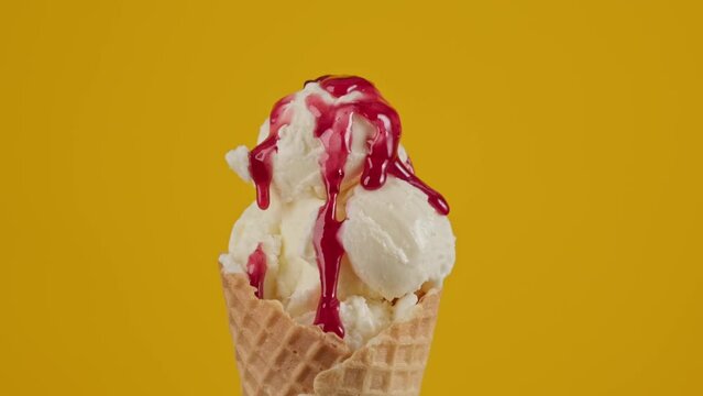 Strawberry topping flowing on white ice cream at yellow background. Ice cream in cone with red sauce and colorful sprinkles. Food concept. Close-up in 4K, UHD