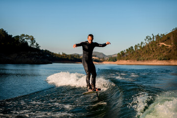 man in wetsuit energetically balancing on river water on a foil wakeboard on beautiful landscape background.