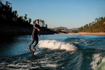handsome man in wetsuit balancing on river water on a foil wakeboard on landscape background.