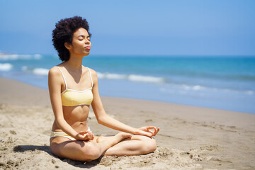 Fototapeta na wymiar Black woman sitting on the beach doing mindfulness meditation in lotus position.