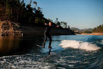 active man in wetsuit energetically balancing on river water on a foil wakeboard on beautiful landscape background.