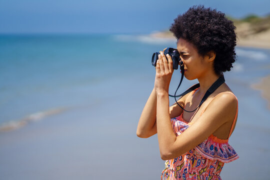 Young black woman with afro hair in summer dress, taking pictures with an SLR camera on the beach.