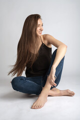 Beauty, fashion and make-up concept. Portrait of happy, beautiful, slim, tall and sexy looking woman with long dark hair wearing black blouse and blue jeans sitting on white studio background floor