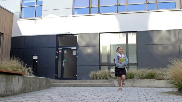 An elementary school student rushes home after school. A little girl with pigtails and a backpack runs against the background of the school building.