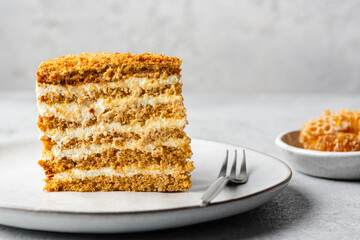 Cut of honey cake with sour cream served on a plate with a fork and honeycombs on a gray concrete background with space for text. Traditional Russian Medovik.