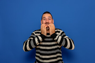 Latino man with his hands on his mouth feeling authentic terror, he bites his nails to try to calm his fears. Isolated on blue studio background.