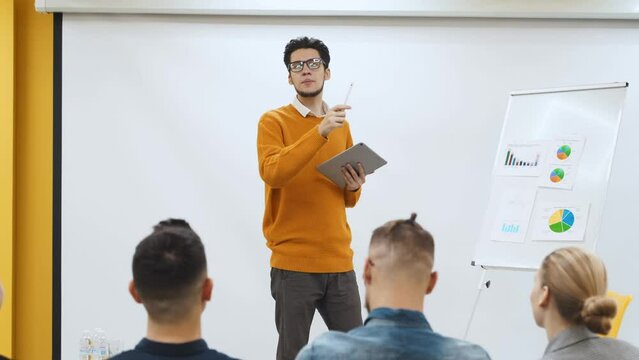 Businessman coach trainer presents report, shows diagrams on flip chart in conference hall. Audience raises hands for answer on presentation in office. Teacher leading master class to audience.