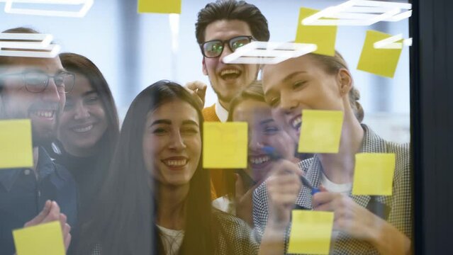 Business team have fun brainstorming and writing ideas on paper sticky notes on a glass wall. Startup team discuss project development plan at scrum meeting. Colleagues find a solution and rejoice.