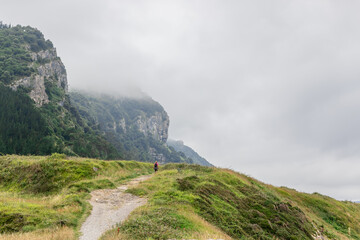 Lone traveler wanders along rocks of Cabo Antzoriz overgrown with emerald grass and moss on foggy summer morning. Lequeitio, Biscay, Basque Country