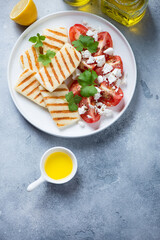 White plate with grilled halloumi, tomatoes and feta, flat lay on a light-blue stone background, vertical shot with space