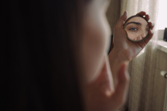 Woman looking in small pocket mirror and applying facial cream on smooth skin during morning beauty routine at home 