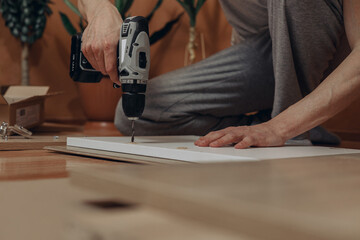 Ground level of crop man drilling hole in wooden board with electric screwdriver while assembling new furniture and sitting on floor in room at home 
