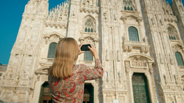 Woman takes Photo, Pictures or Video by Smart Phone in Milan, Italy with Landmark Cathedral Basilica on sunny summer day. Tourist Lady Travels in Europe. 4K backside view low angle orbit shot