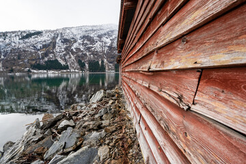 Facade of an old wooden building of Scandinavian type with red wooden boards, where there is a stone wall at the bottom behind which you can see the water