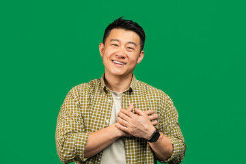 Positive middle aged asian man praying and smiling at camera, holding hands on his chest over...
