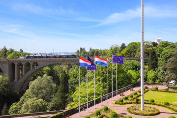 View of the Adolphe Bridge in Luxembourg City. Flags of Luxembourg and the European Union fly in the foreground - obrazy, fototapety, plakaty