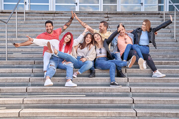 Obraz premium Group of happy young friends having fun and smiling at camera while sitting together on stairs outdoors. Friendship and positivity concept.